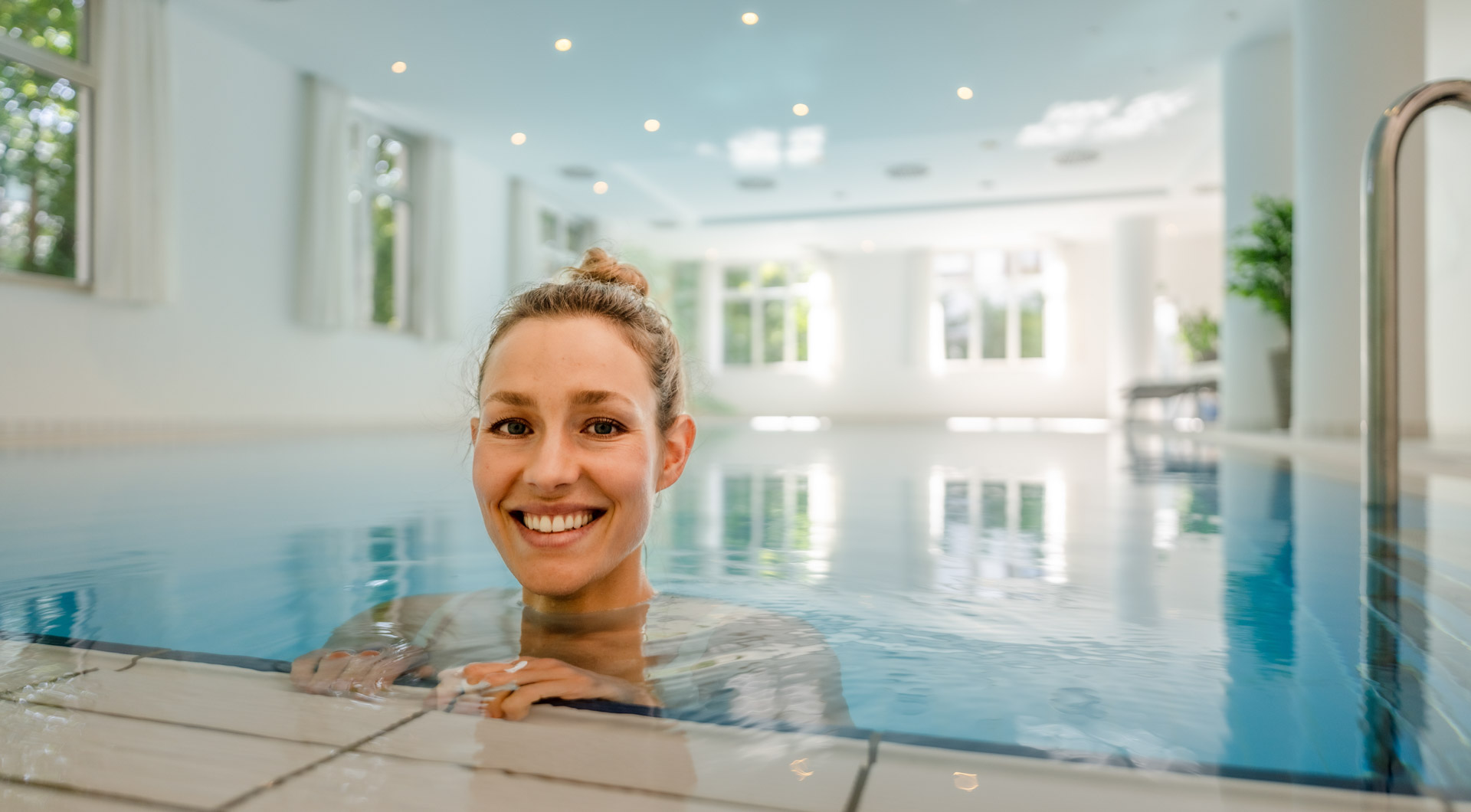 Weiblicher Gast, welcher im Pool am Beckenrand in die Kamera lächelt.